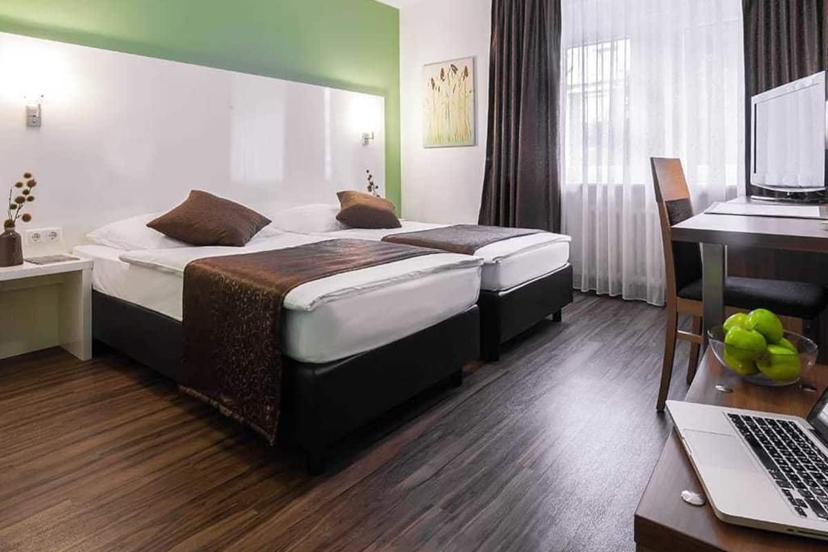 A hotel room with two beds, a desk with a laptop, a chair, and a window with curtains.