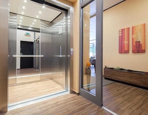 A modern elevator with a glass door, a wooden floor, and a wall with abstract paintings. - Hotel Mondial (Langenfeld)