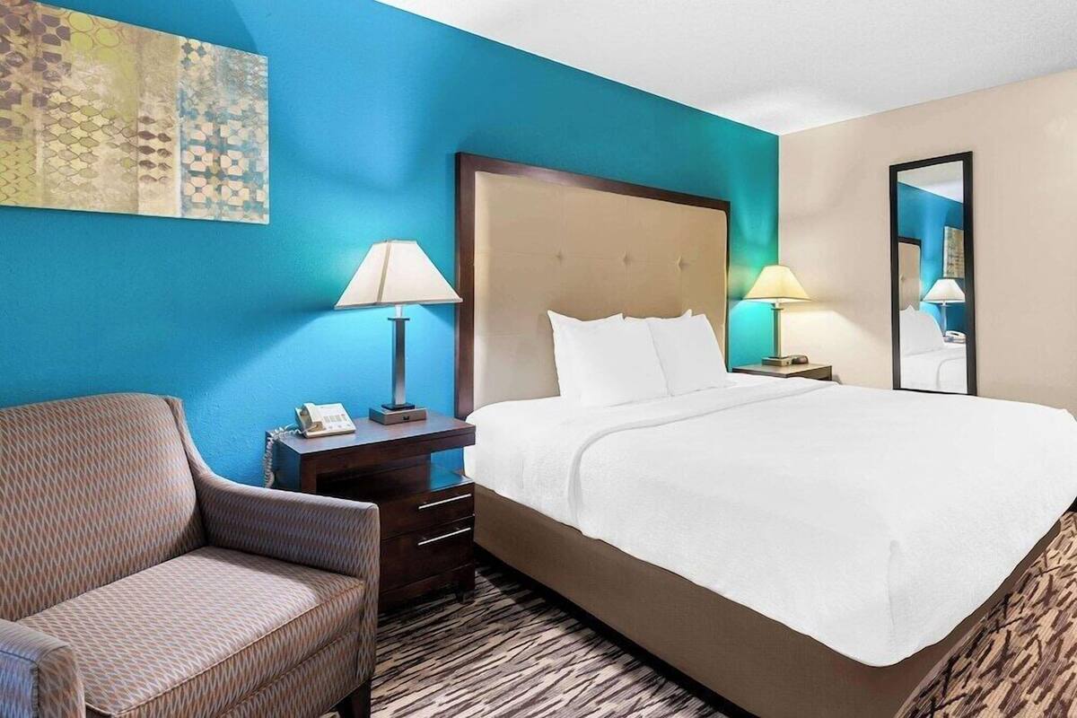 A hotel room with a large bed, a chair, a desk with a lamp, and a mirror.