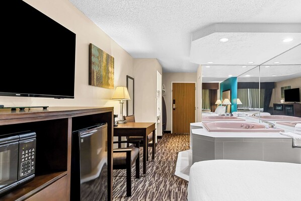 A hotel room with a flat-screen TV, a dining table with chairs, a kitchenette with a microwave, and a large bathroom with a bathtub and sink.