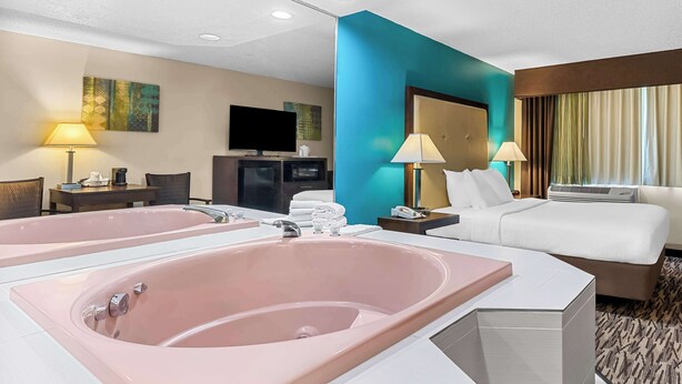A hotel room with a large pink bathtub, a bed, a desk with a lamp, a television, and a window with curtains.