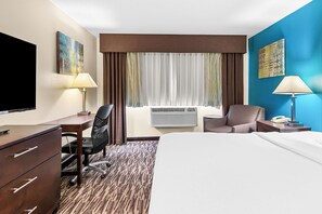 A hotel room with a bed, desk, chair, TV, air conditioning unit, and artwork on the walls.