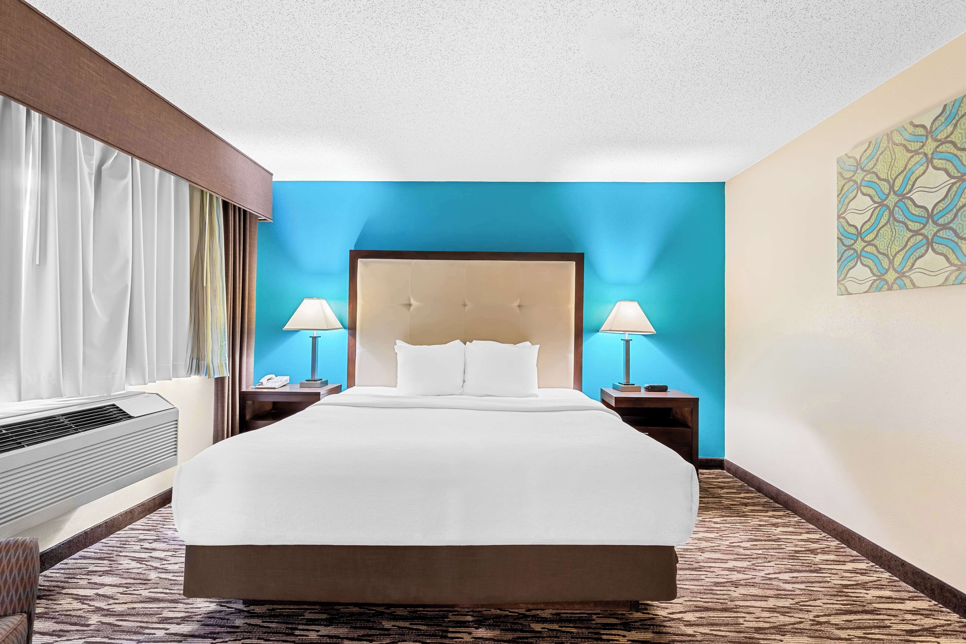 A hotel room with a large bed, two bedside tables with lamps, a wall-mounted air conditioner, and a patterned carpet.