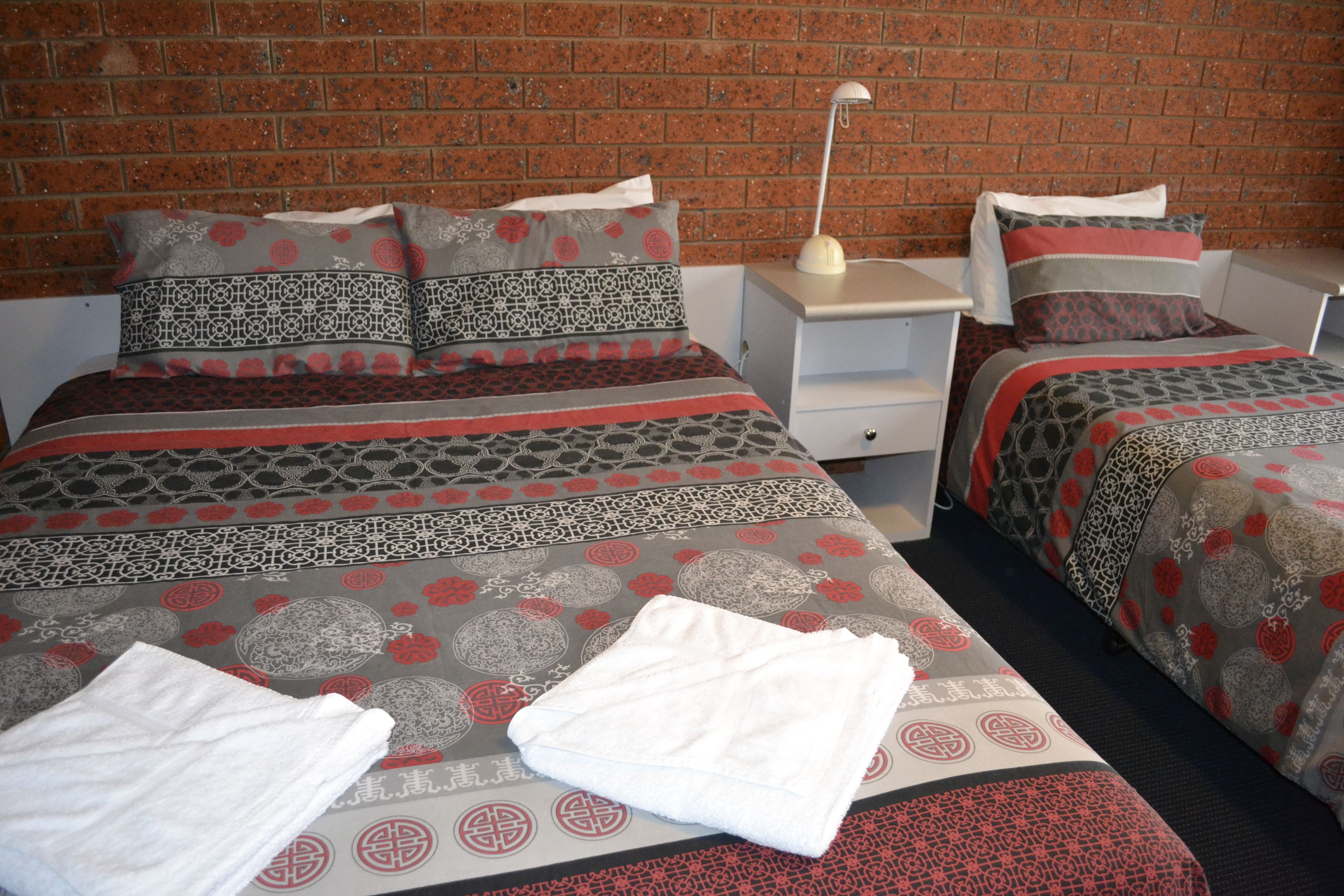 A hotel room with two beds, each with patterned bedding and pillows, a nightstand with a lamp, and a brick wall.