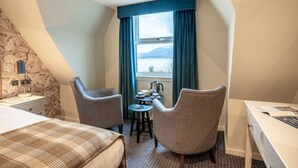 A hotel room with a bed, two armchairs, a small table, and a view of a lake and mountains through a window. - Ballachulish Hotel (Ballachulish)