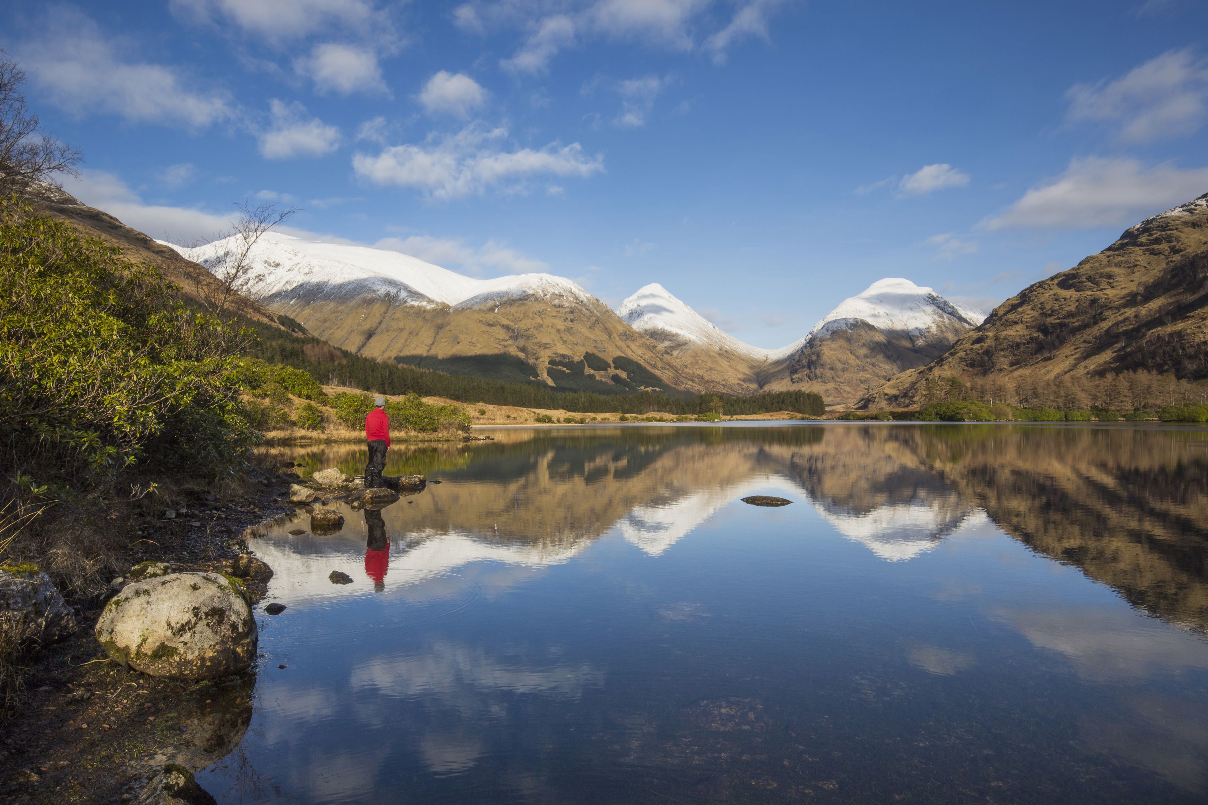 A calm lake with mountains in the background and a person in a red jacket standing by the shore.