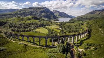A large, arched stone bridge in a lush, green valley with a lake and mountains in the background.
