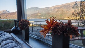 A room with a large window overlooking a lake and mountains, a cushioned bench, and two vases with dried plants. - Ballachulish Hotel (Ballachulish)