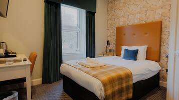 A hotel room with a bed, a desk, a chair, a window with curtains, and a patterned wall.