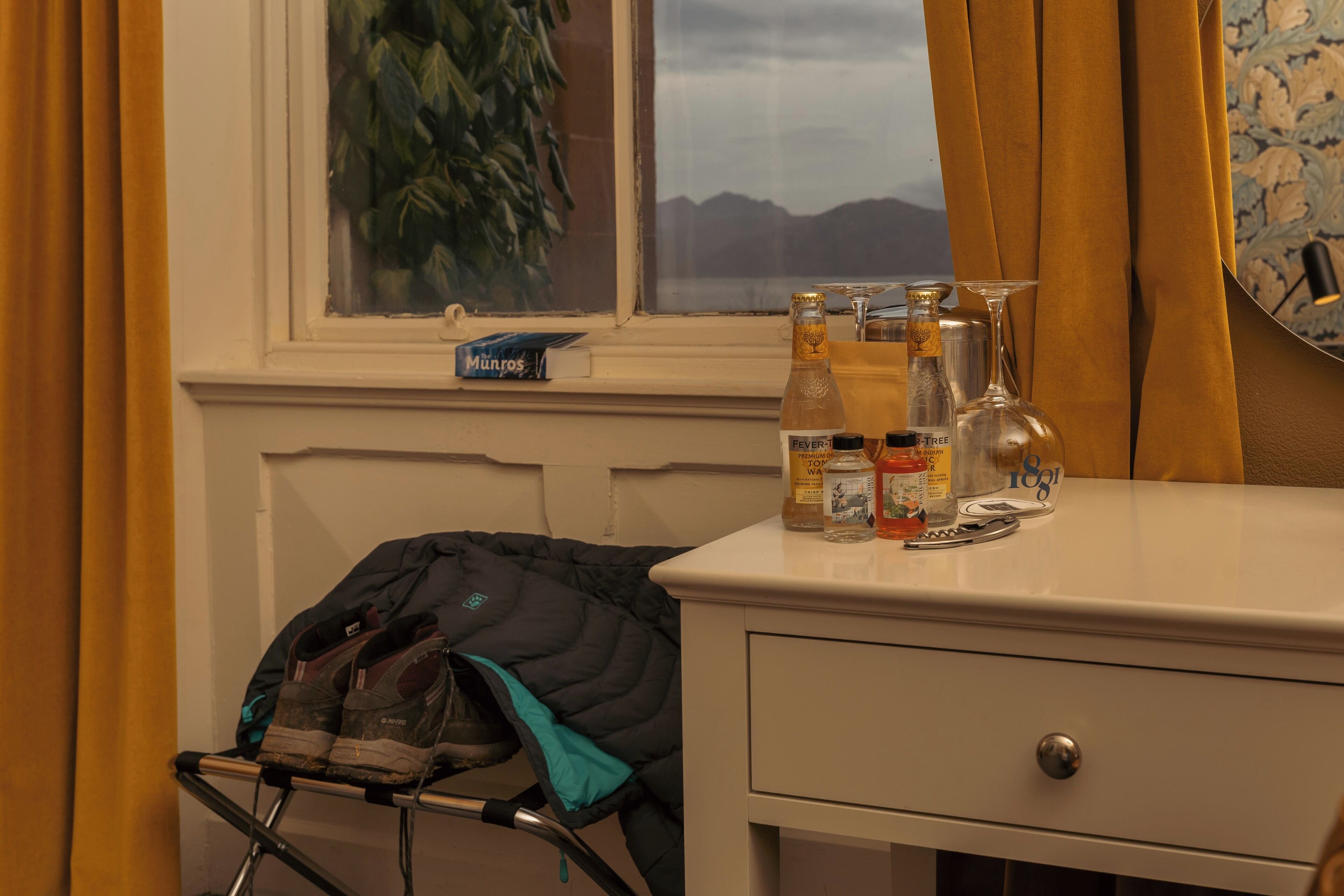 A hotel room with a window, a desk with bottles, a jacket on a rack, and a plant.