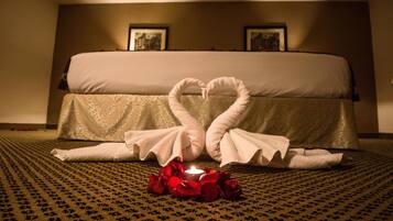 A hotel room with a bed, a heart-shaped towel arrangement, and a lit candle surrounded by rose petals.