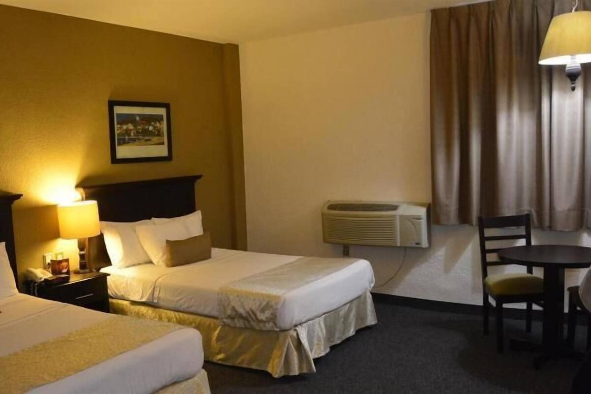 A hotel room with two beds, a desk, a chair, a lamp, an air conditioner, and two framed pictures on the wall.