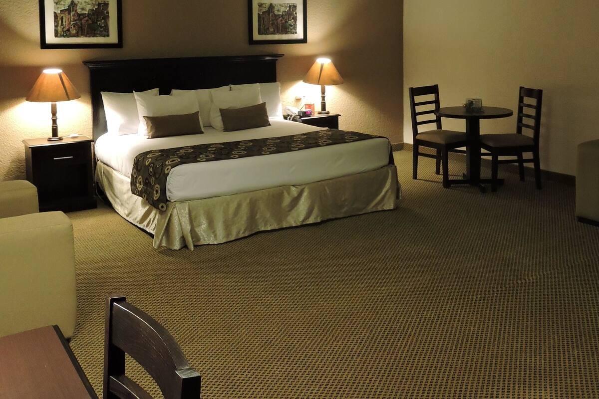 A hotel room with a large bed, two bedside lamps, a small table with chairs, and two framed pictures on the wall.
