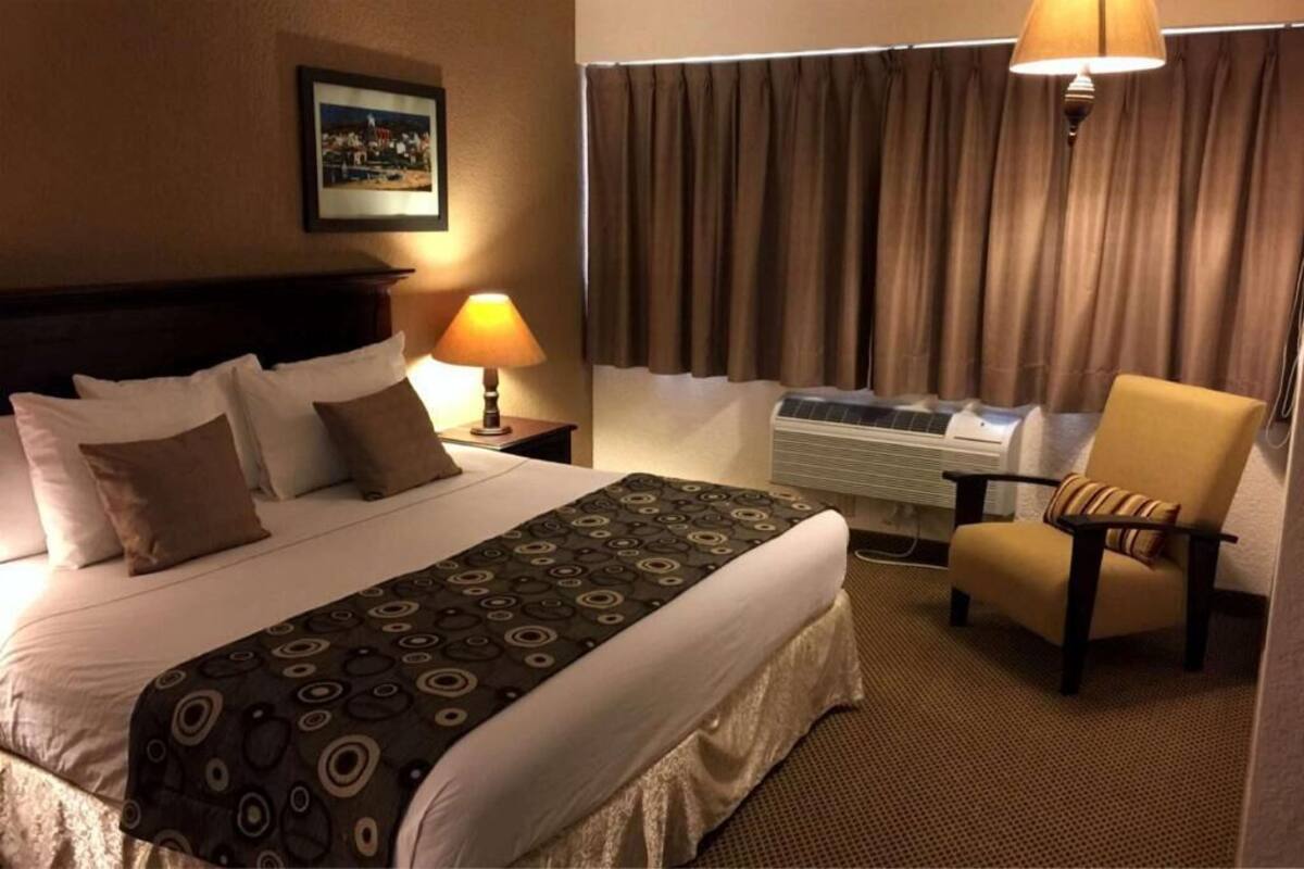A hotel room with a bed, a chair, a lamp, curtains, and an air conditioning unit.