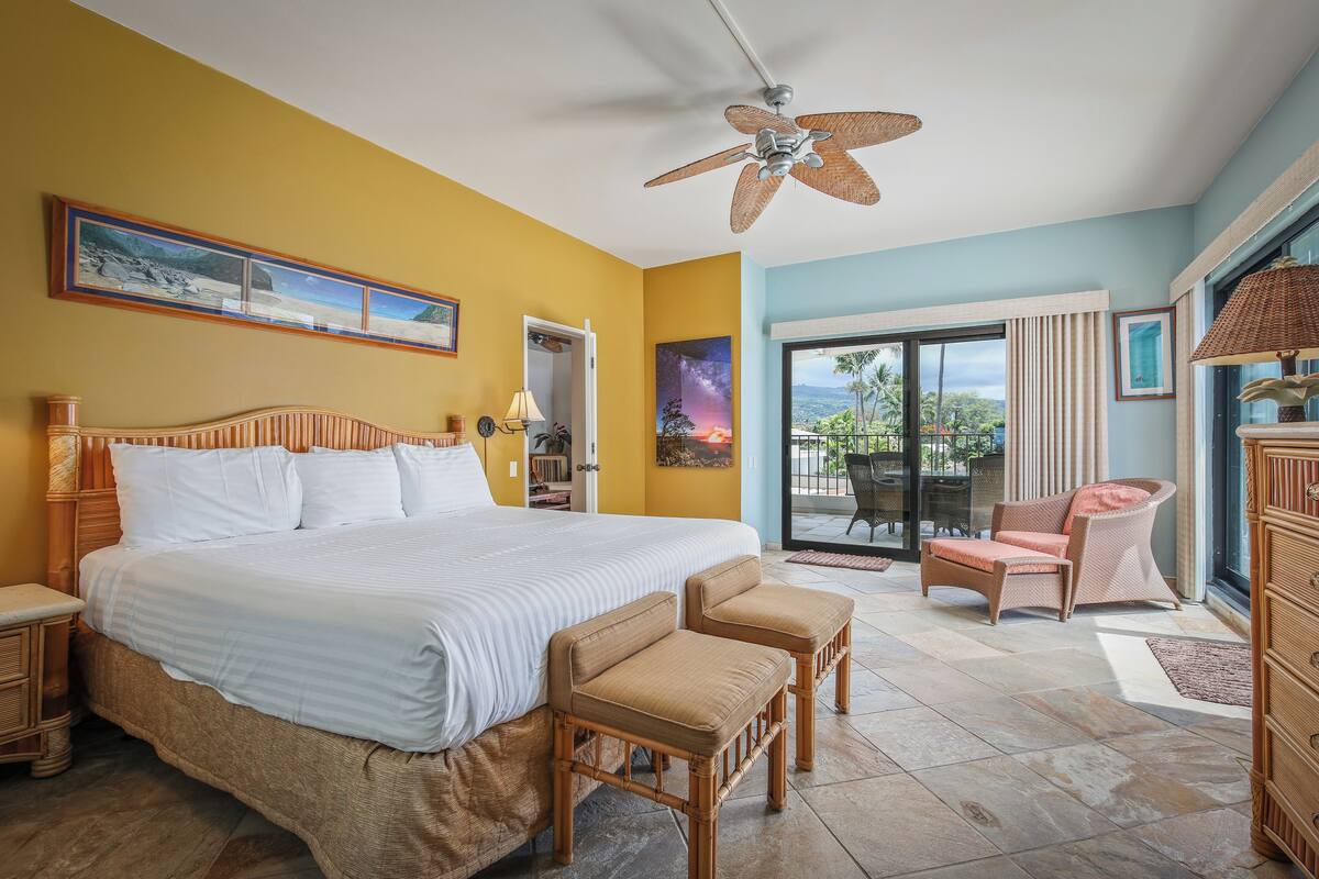 Une chambre avec un grand lit, deux fauteuils, un ventilateur de plafond et une vue sur l’extérieur.