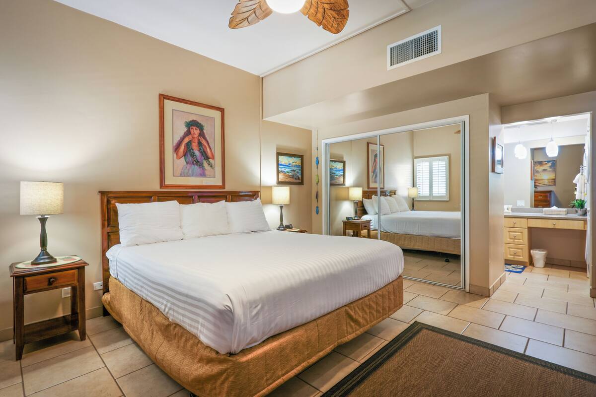 Une chambre d’hôtel avec un grand lit, une table de chevet avec une lampe, un miroir et une salle de bain visible à travers une porte ouverte.