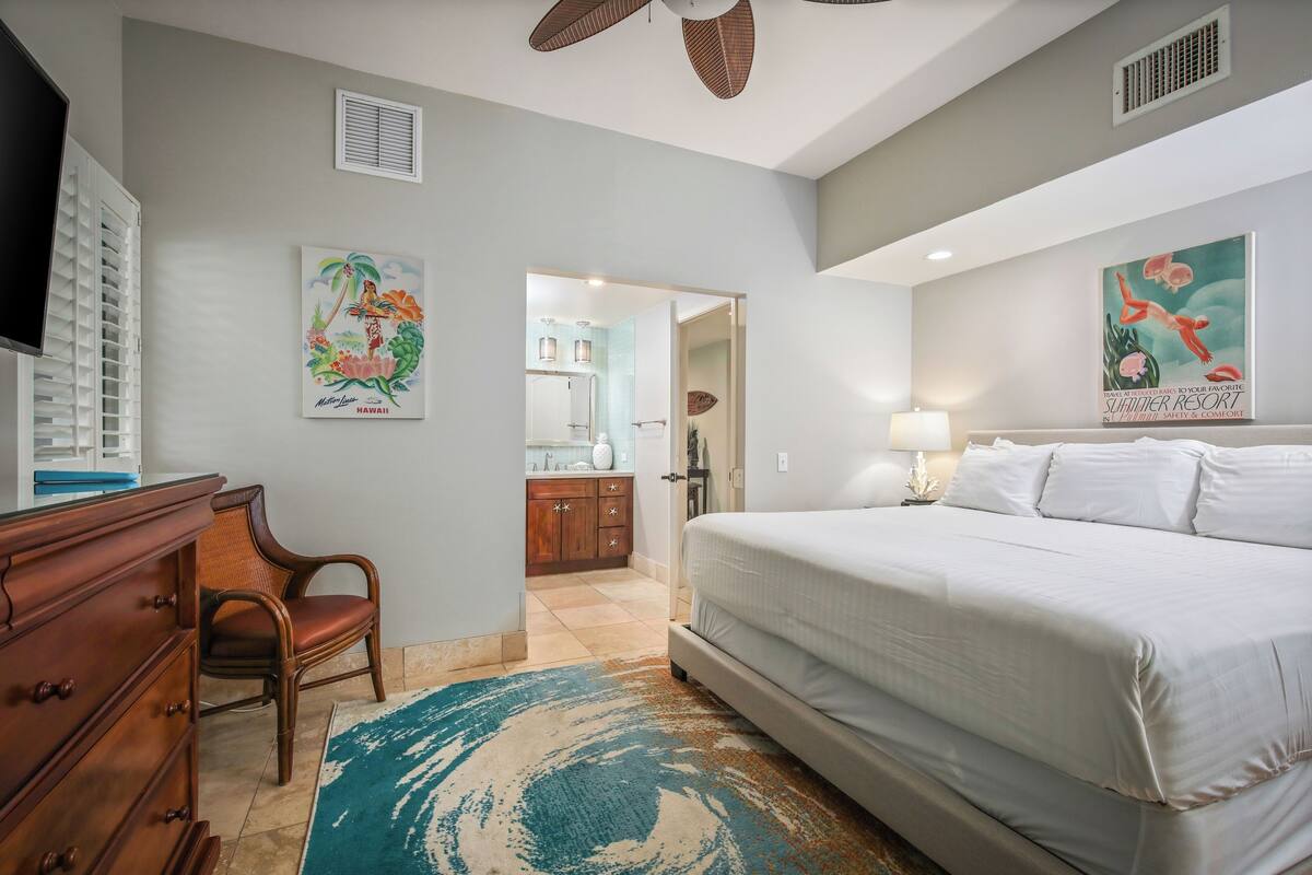 Une chambre à coucher avec un lit, une commode, une chaise et une salle de bain visible à travers une porte ouverte.