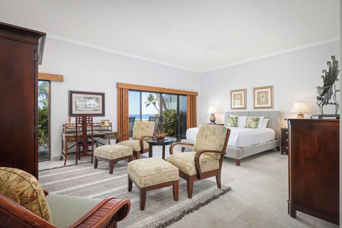Une chambre d’hôtel avec un lit, deux fauteuils, une table à manger et un bureau. Des tableaux encadrés ornent le mur et une lampe se trouve sur le bureau.