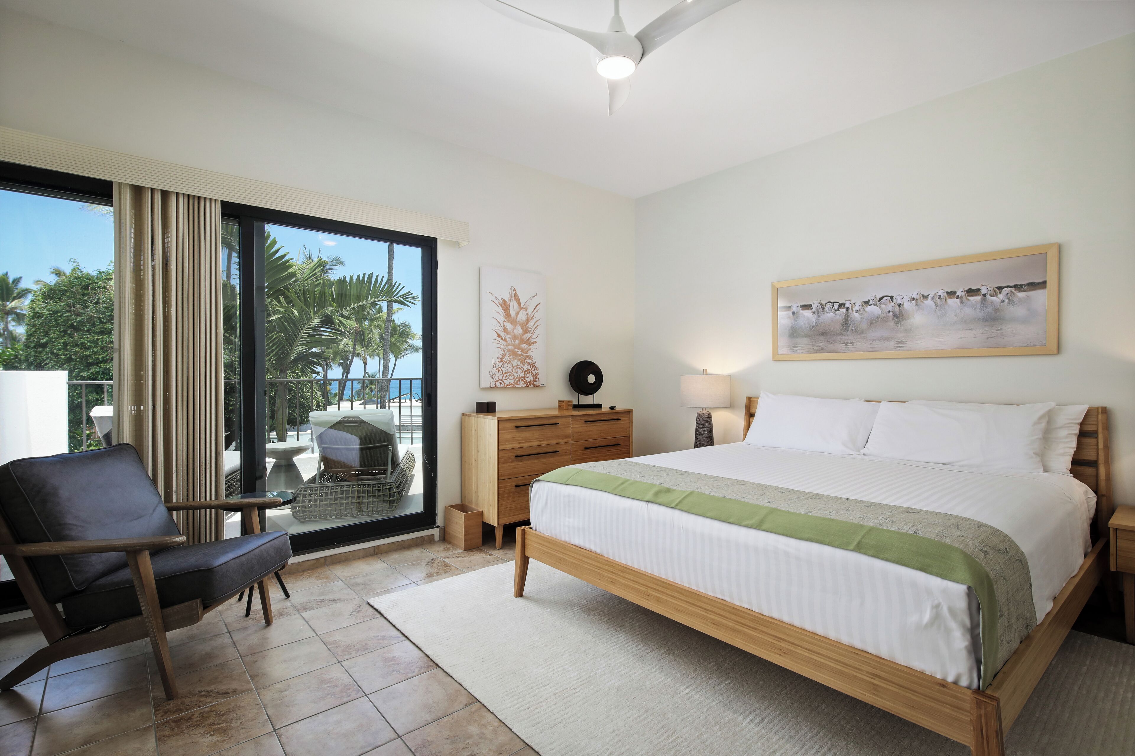 Habitación de hotel con cama, una silla, un escritorio, una lámpara, un cuadro y vistas a palmeras a través de un ventanal grande.