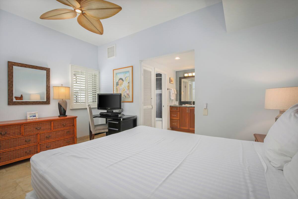 Une chambre d’hôtel avec un grand lit, un bureau avec un ordinateur, une commode, une chaise et un ventilateur de plafond.