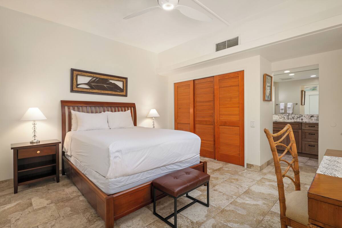 Une chambre à coucher bien rangée, avec un lit, des tables de chevet, un banc, une chaise, un ventilateur de plafond et une armoire en bois.