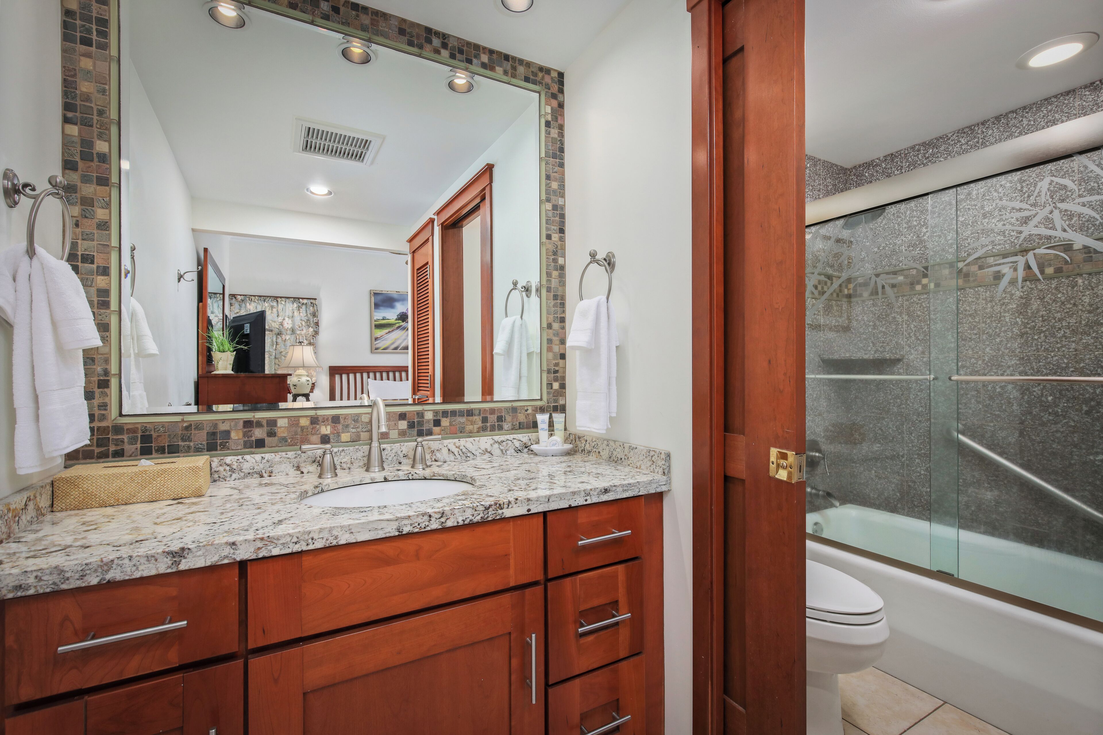 A bathroom with a granite countertop, wooden cabinets, a large mirror, and a glass-enclosed shower.