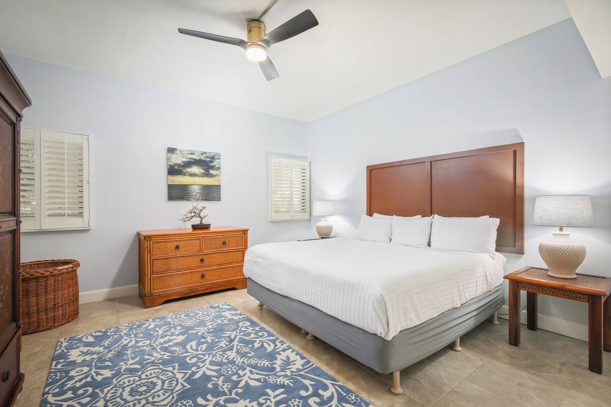 Une chambre à coucher comprenant un lit, une table de chevet, une commode, un ventilateur de plafond et une lampe fixée au mur.