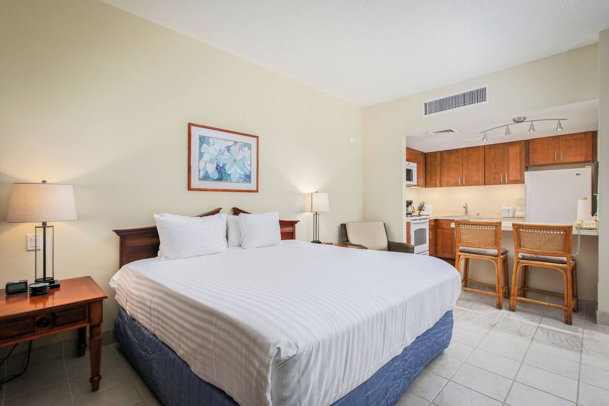 Une chambre d’hôtel avec un grand lit, deux tables de chevet, un bureau, une chaise, un coin cuisine et un tableau au mur.