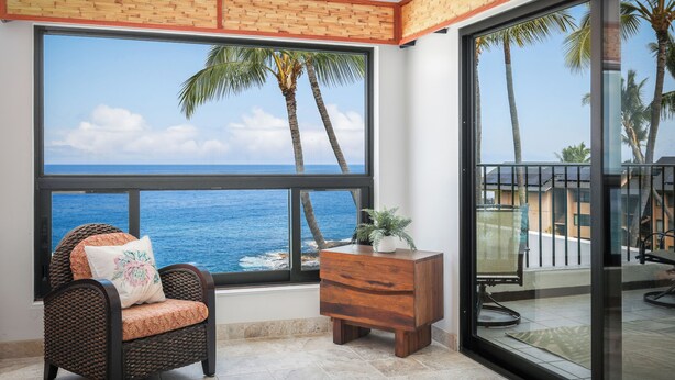 A room with a large window overlooking the ocean, a wicker chair with a cushion, and a wooden side table with a plant.