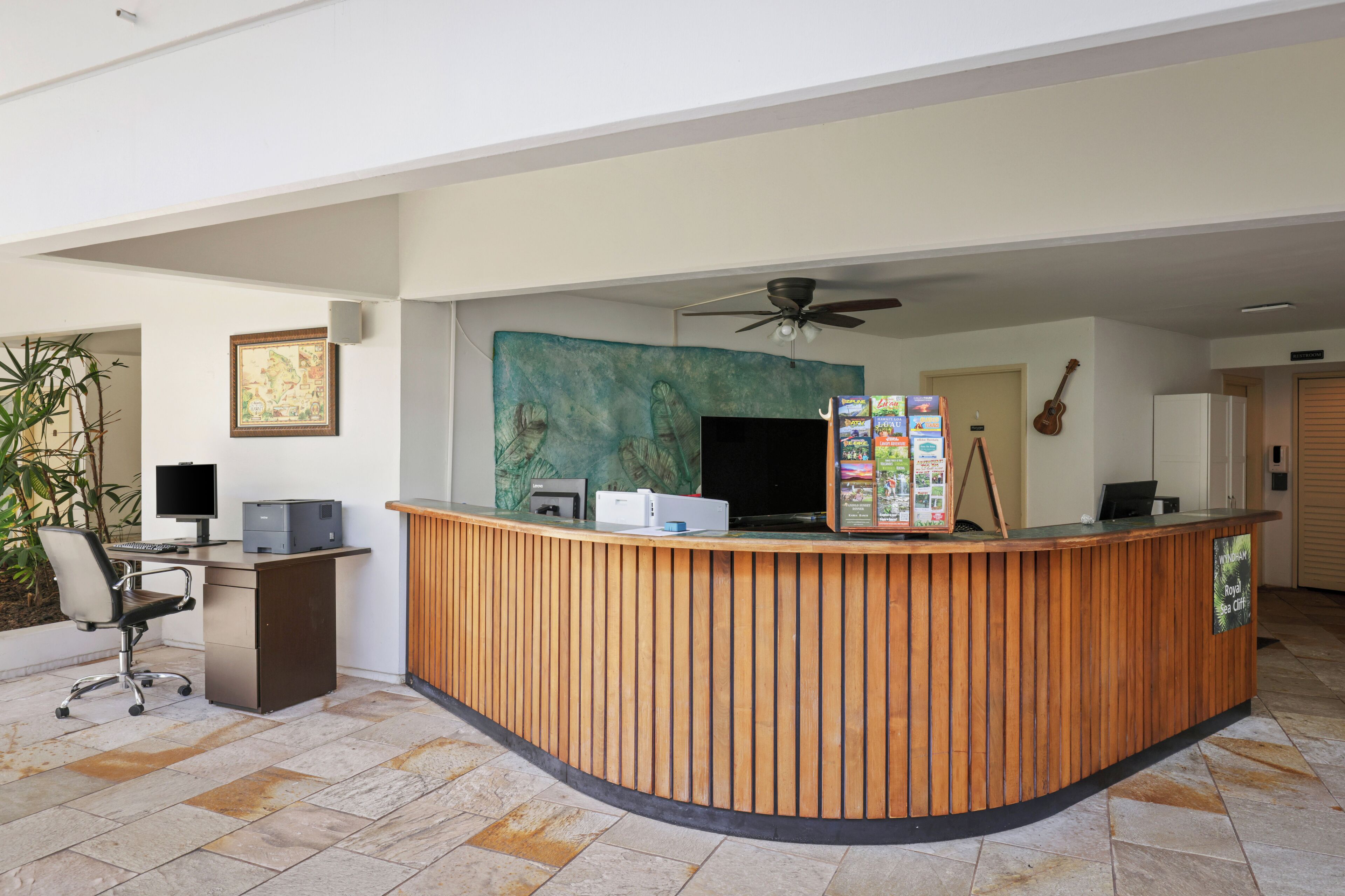 A reception desk with a computer, printer, and a wall-mounted TV. There is a ceiling fan, a guitar, and a framed picture on the wall.