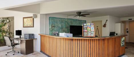 A reception desk with a computer, printer, and a wall-mounted TV. There is a ceiling fan, a guitar, and a framed picture on the wall.