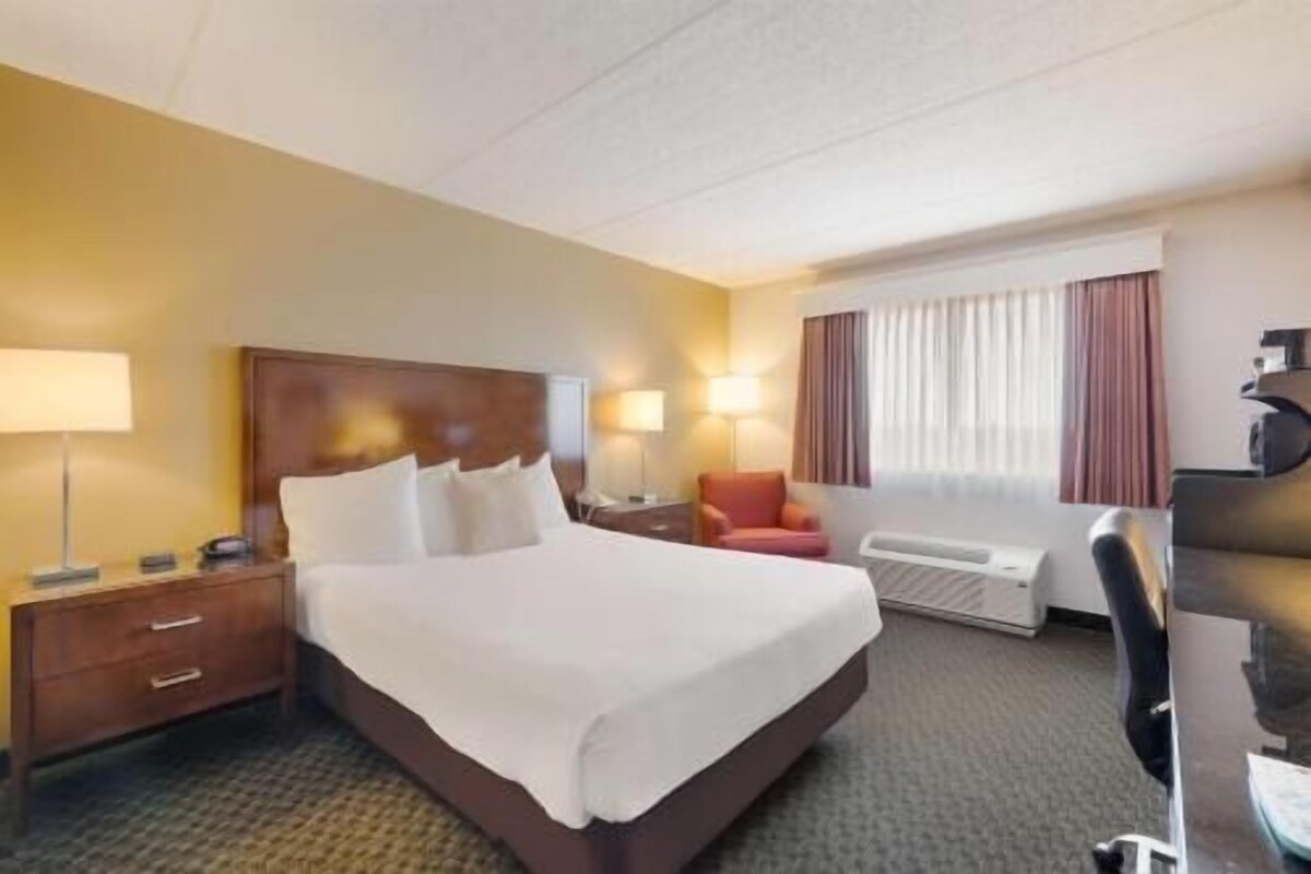 A hotel room with a bed, bedside tables, a desk, a chair, a heater, and a window with curtains.