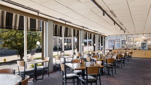 Een restaurant met een lange toonbank, meerdere tafels en stoelen. Er zijn grote ramen langs de muur, waardoor natuurlijk licht en uitzicht op de straat mogelijk zijn.