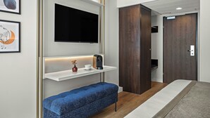 Habitació d'hotel moderna amb televisió de pantalla plana, una taula de nit amb cafetera, una banca blava i un armari.