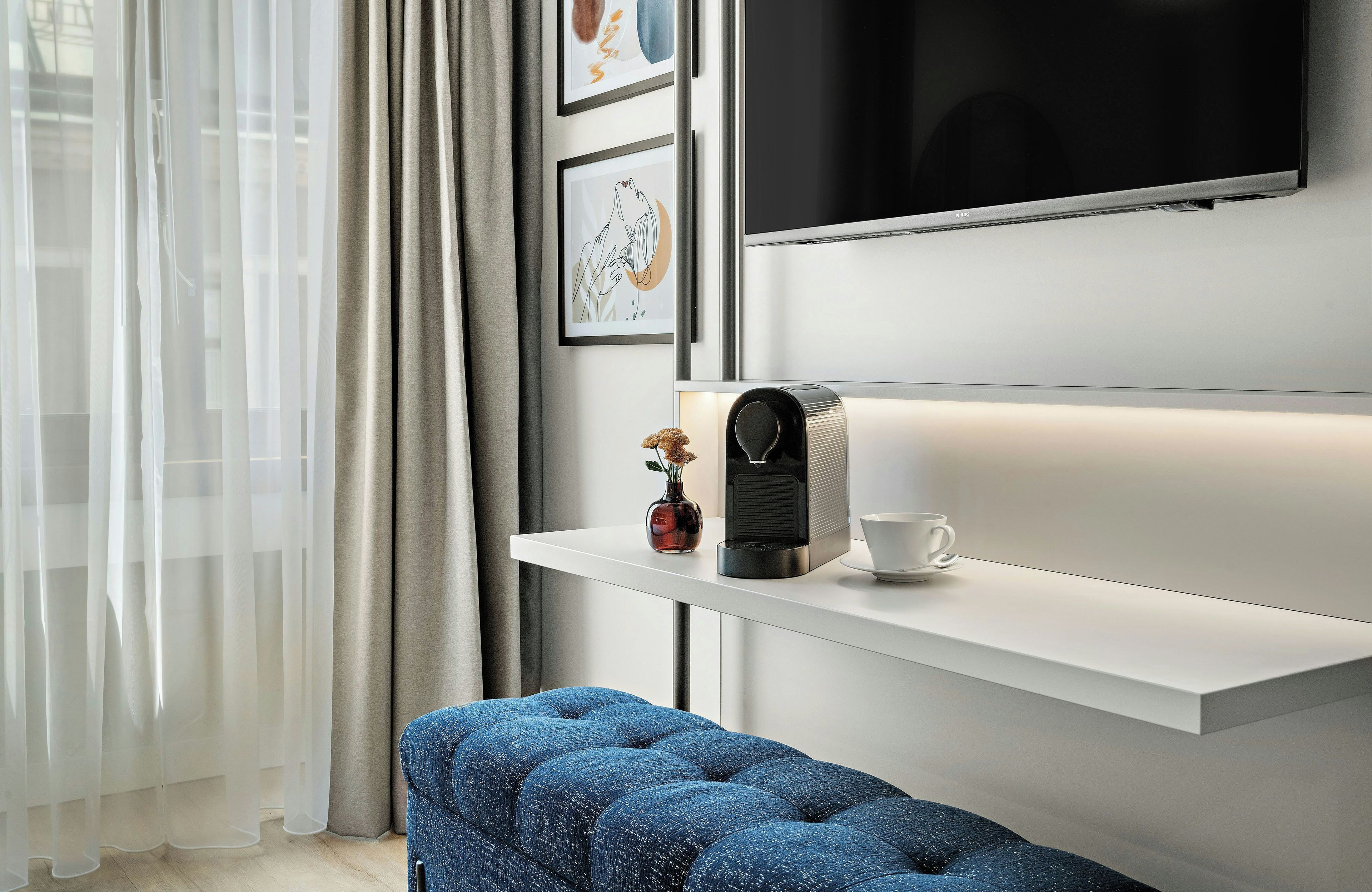 Quarto de hotel moderno com escrivaninha branca, máquina de café, xícara e um banco acolchoado azul.
