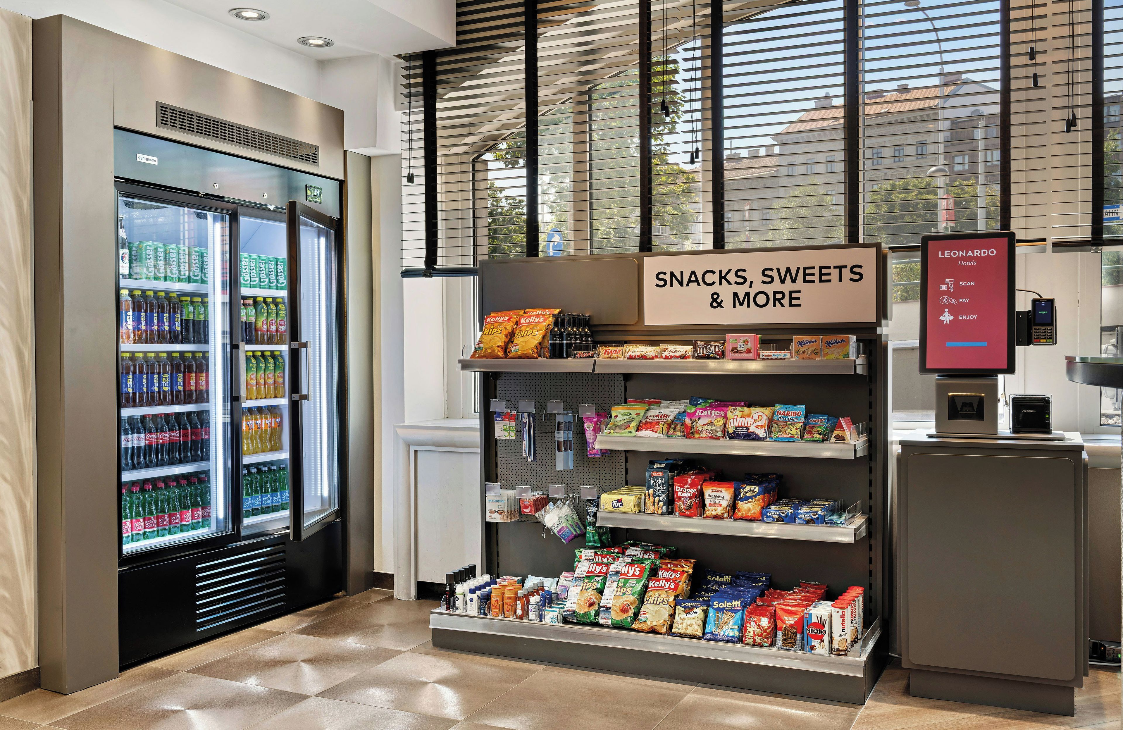 Ein Bereich für Snacks und Getränke im Hotel mit einem Getränkespender, Snackregalen und einem Zahlungsterminal.