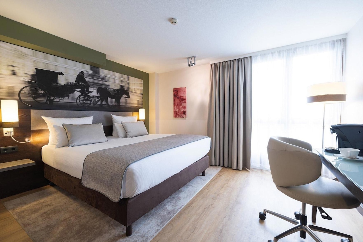 Habitación de hotel con cama, escritorio, silla, lámpara, un cuadro y ventana con cortinas.