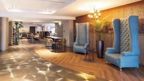 Un lobby moderno con piso de madera curva, una zona de estar con dos sillones azules, una mesita y una planta en maceta.