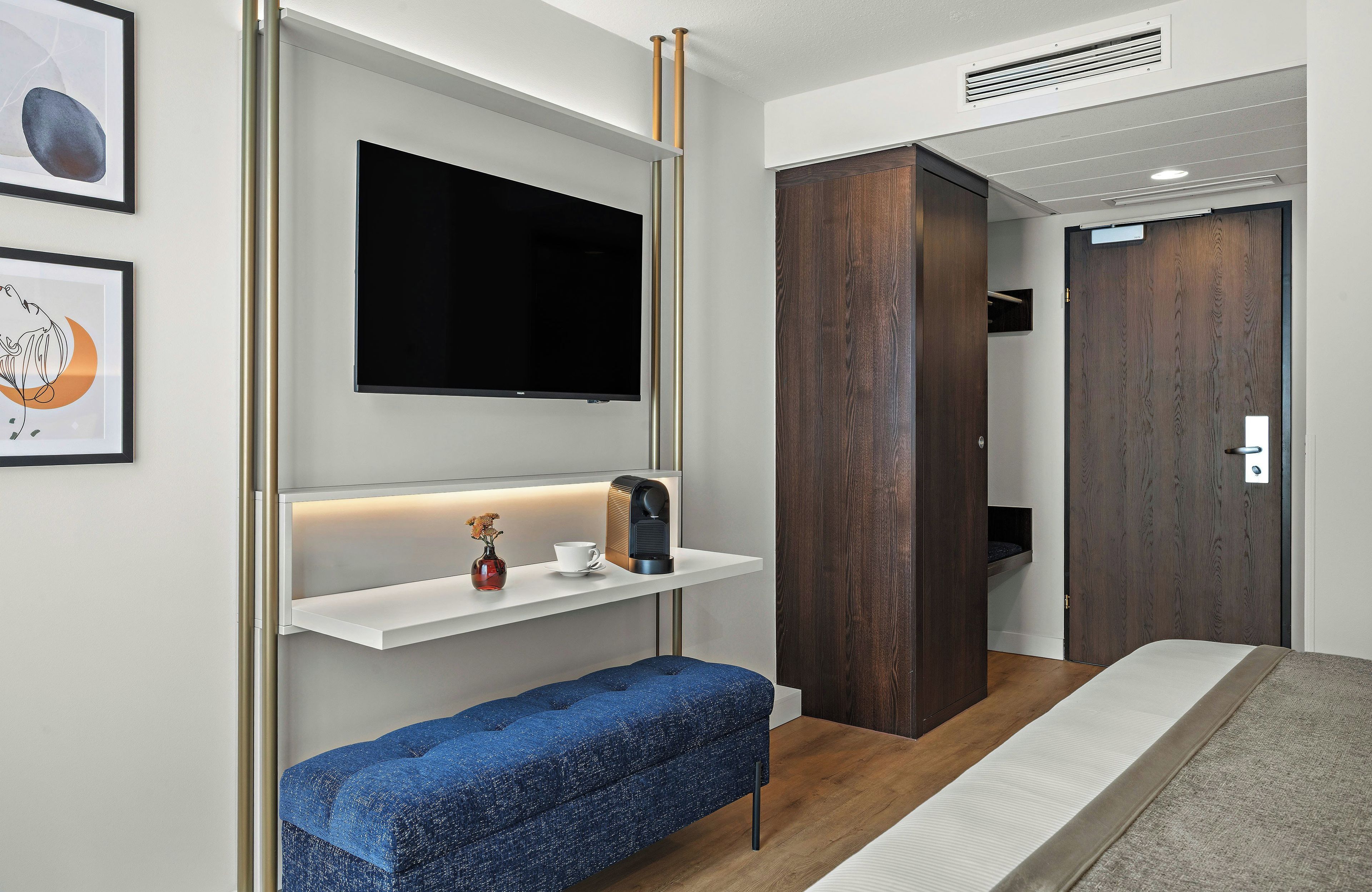 Ein modernes Hotelzimmer mit einem Flachbildfernseher, einem Nachttisch mit Kaffeemaschine, einer blauen Bank und einem Kleiderschrank.