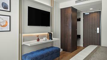 Ein modernes Hotelzimmer mit einem Flachbildfernseher, einem Nachttisch mit Kaffeemaschine, einer blauen Bank und einem Kleiderschrank.