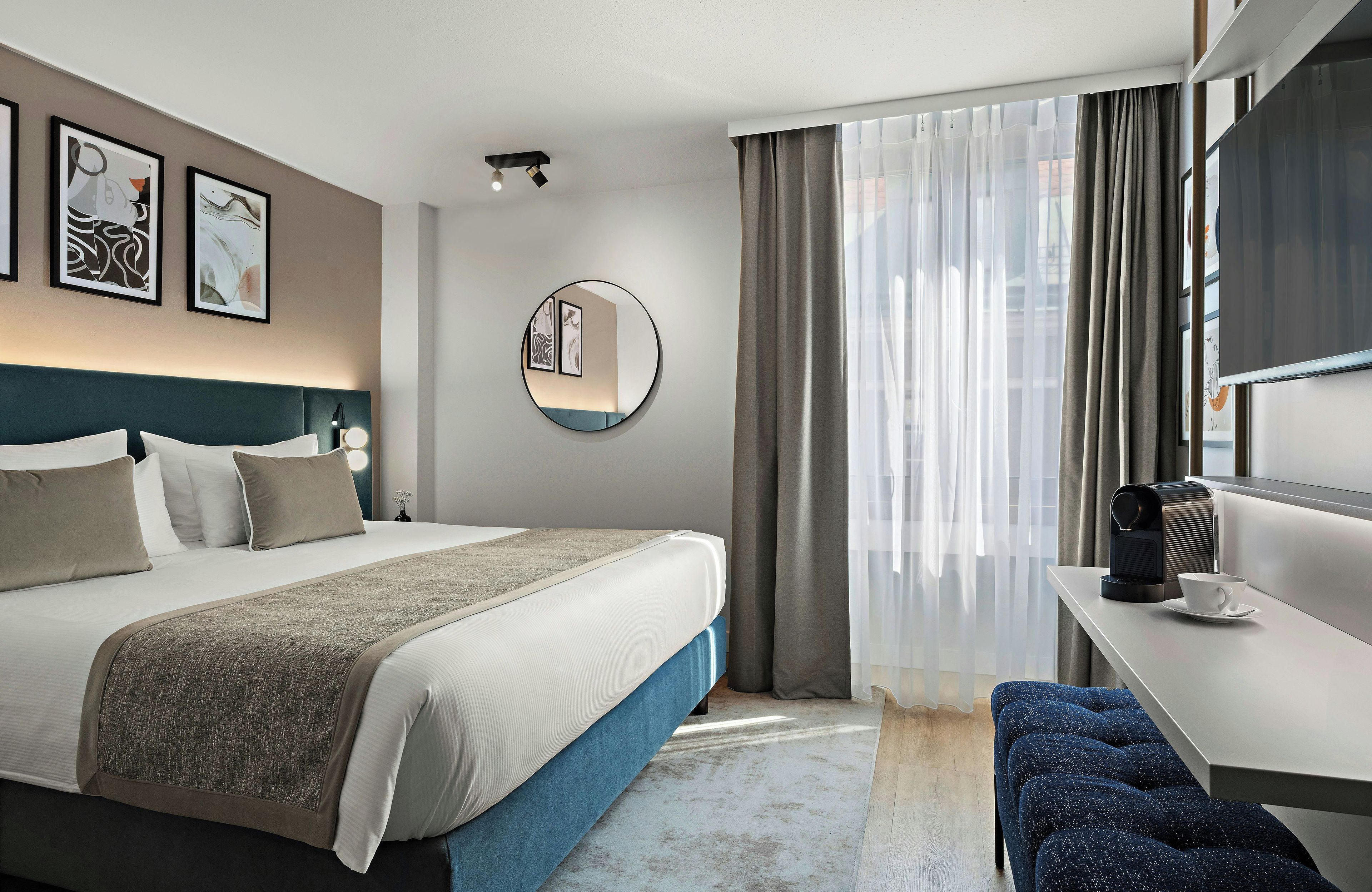 Ein modernes Hotelzimmer mit einem ordentlich bezogenen Bett, einem runden Spiegel, einem Schreibtisch mit Kaffeemaschine und Blick durch leichte Vorhänge.