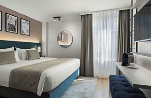 A modern hotel room with a neatly made bed, a round mirror, a desk with a coffee maker, and a view through sheer curtains. - Leonardo Hotel Vienna Westbahnhof (Vienna)