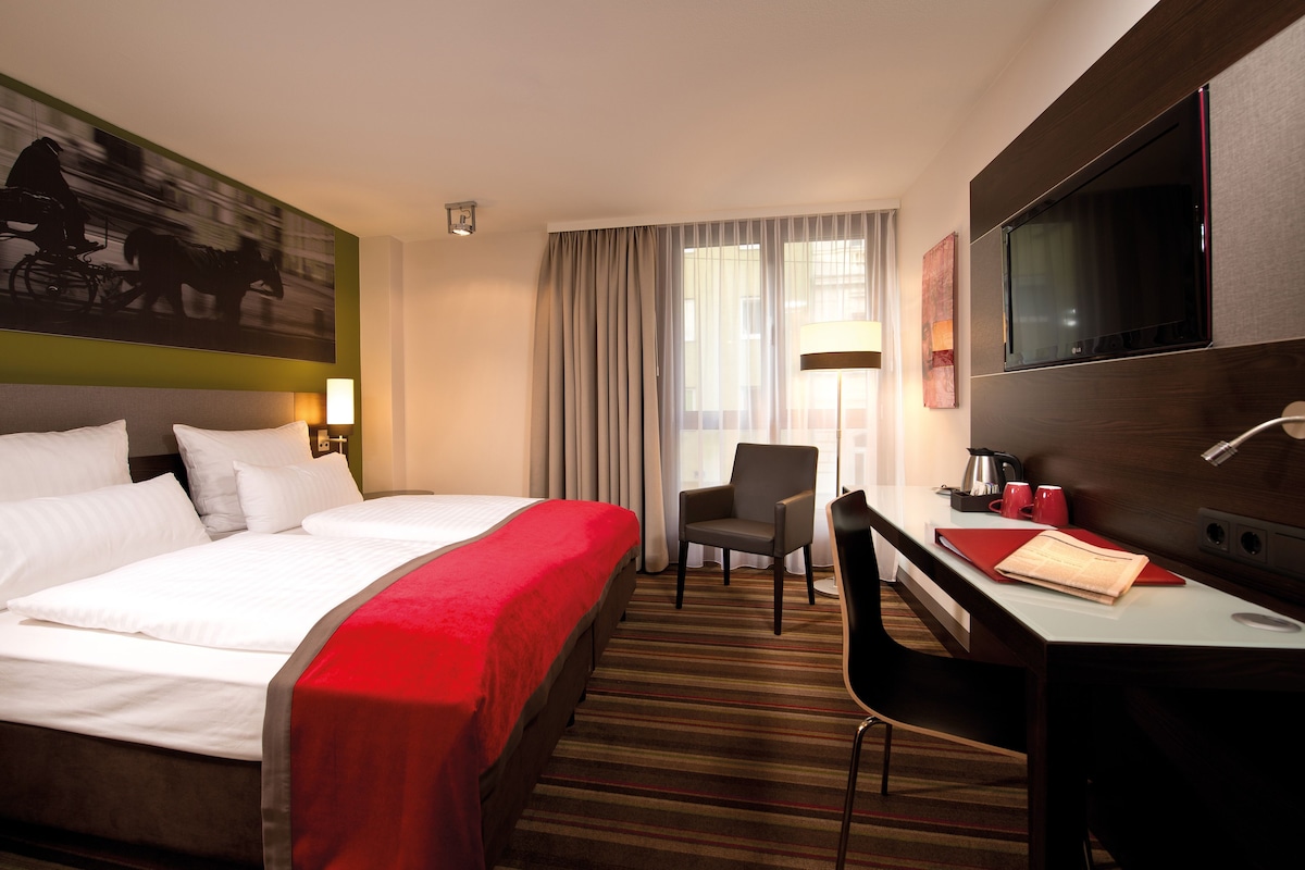 Habitación de hotel con cama, escritorio, silla, televisor y cortinas.