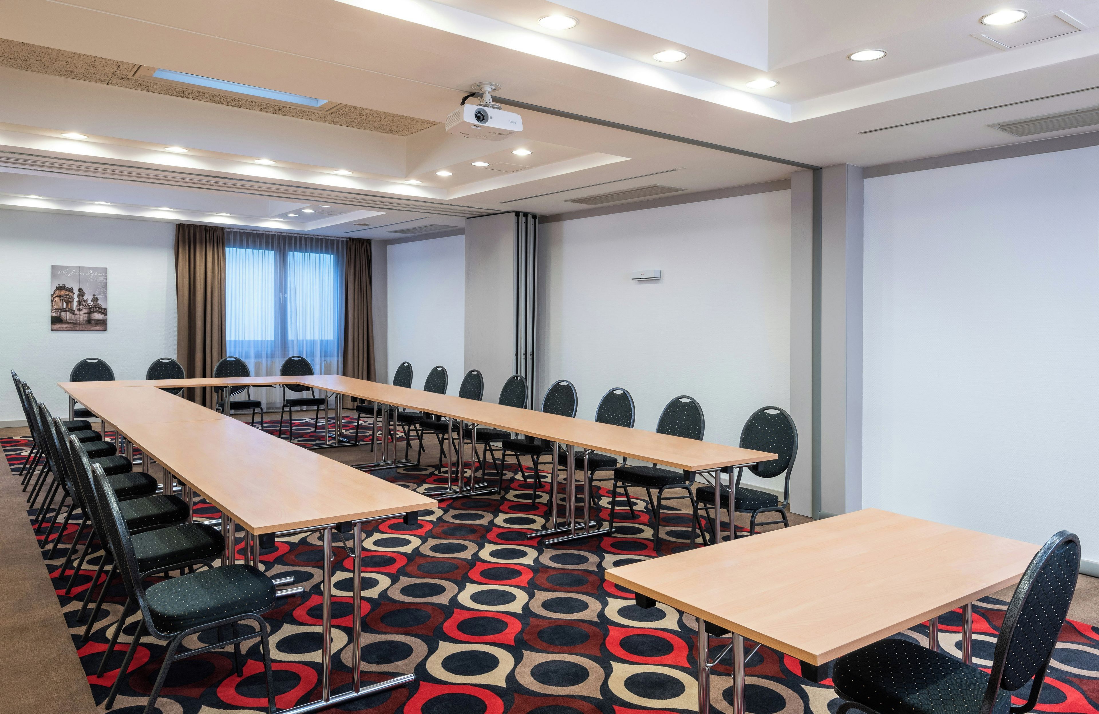 Une salle de conférence avec de longues tables, des chaises, un projecteur et une moquette à motifs.