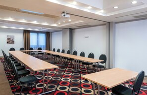 Ein Konferenzraum mit langen Tischen, Stühlen, einem Projektor und einem gemusterten Teppich.