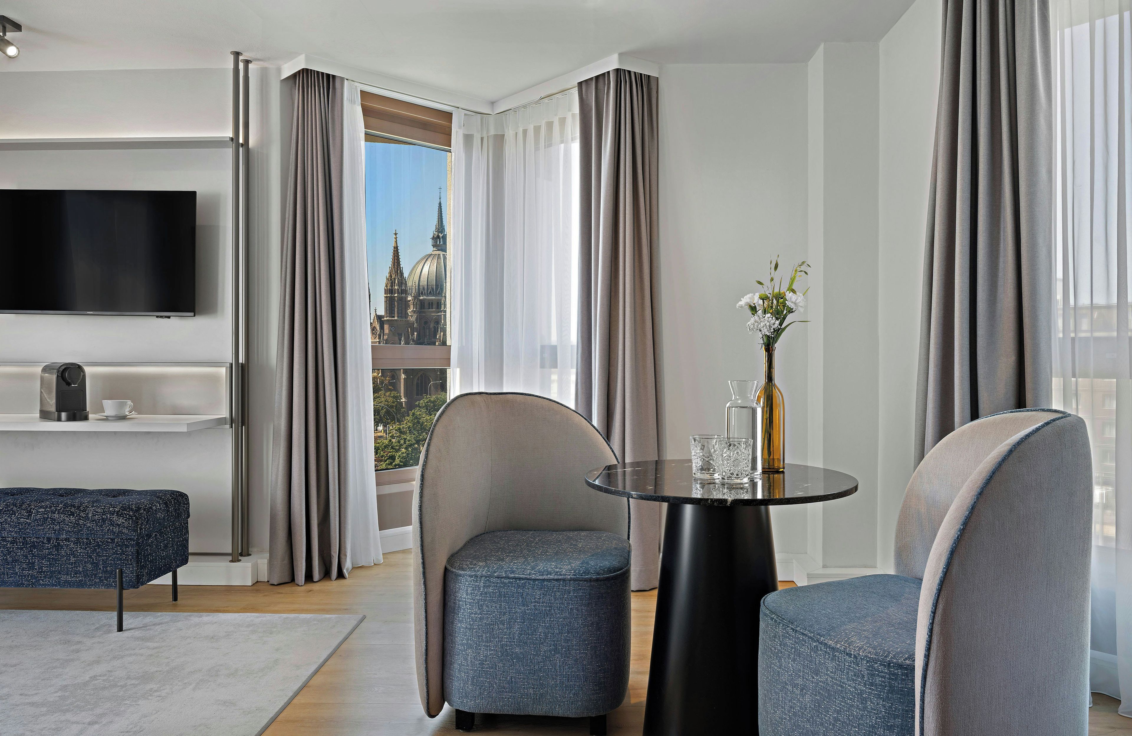 Une chambre d’hôtel moderne dotée d’une grande fenêtre, d’un téléviseur à écran plat, d’une table ronde avec des chaises et d’un petit canapé.