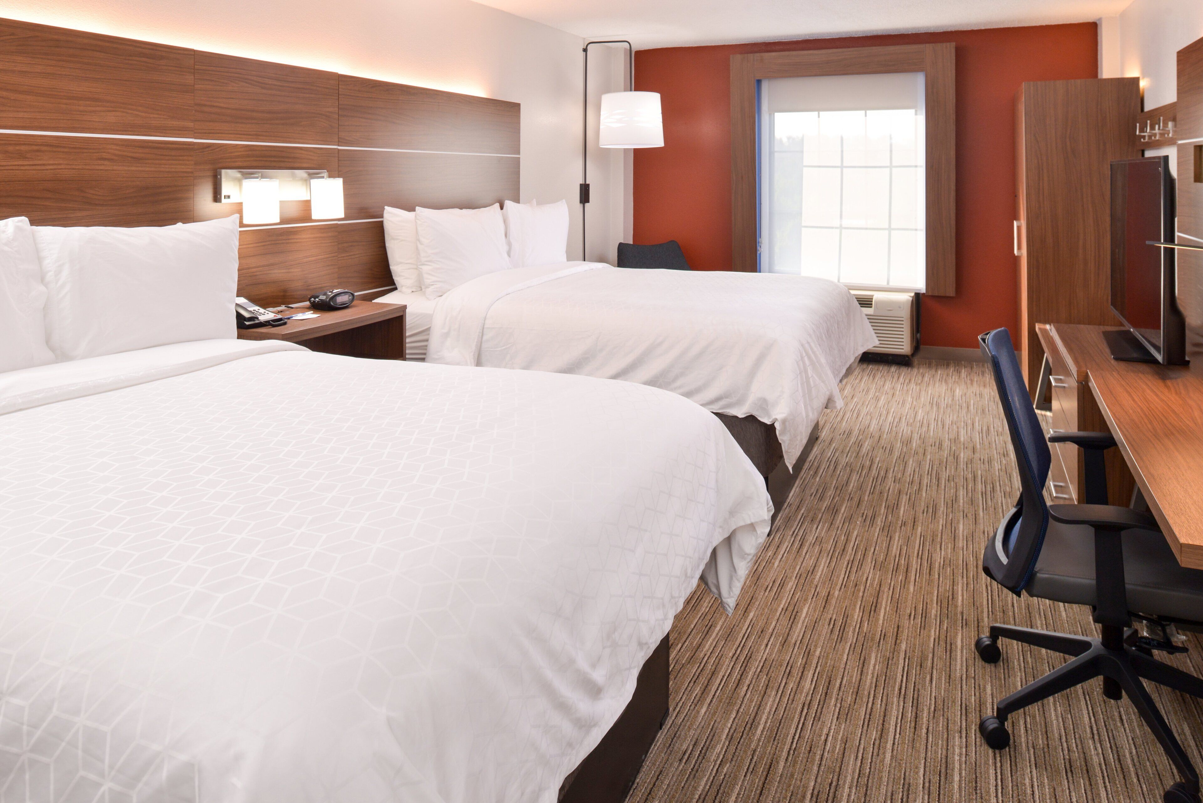 A hotel room with two beds, a desk, a chair, a lamp, and a window with blinds.