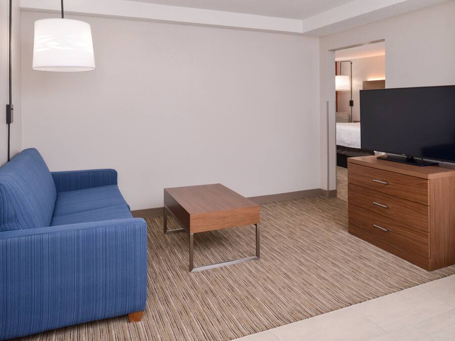 A modern hotel room with a blue sofa, a wooden coffee table, a flat-screen TV on a wooden cabinet, and a large window.