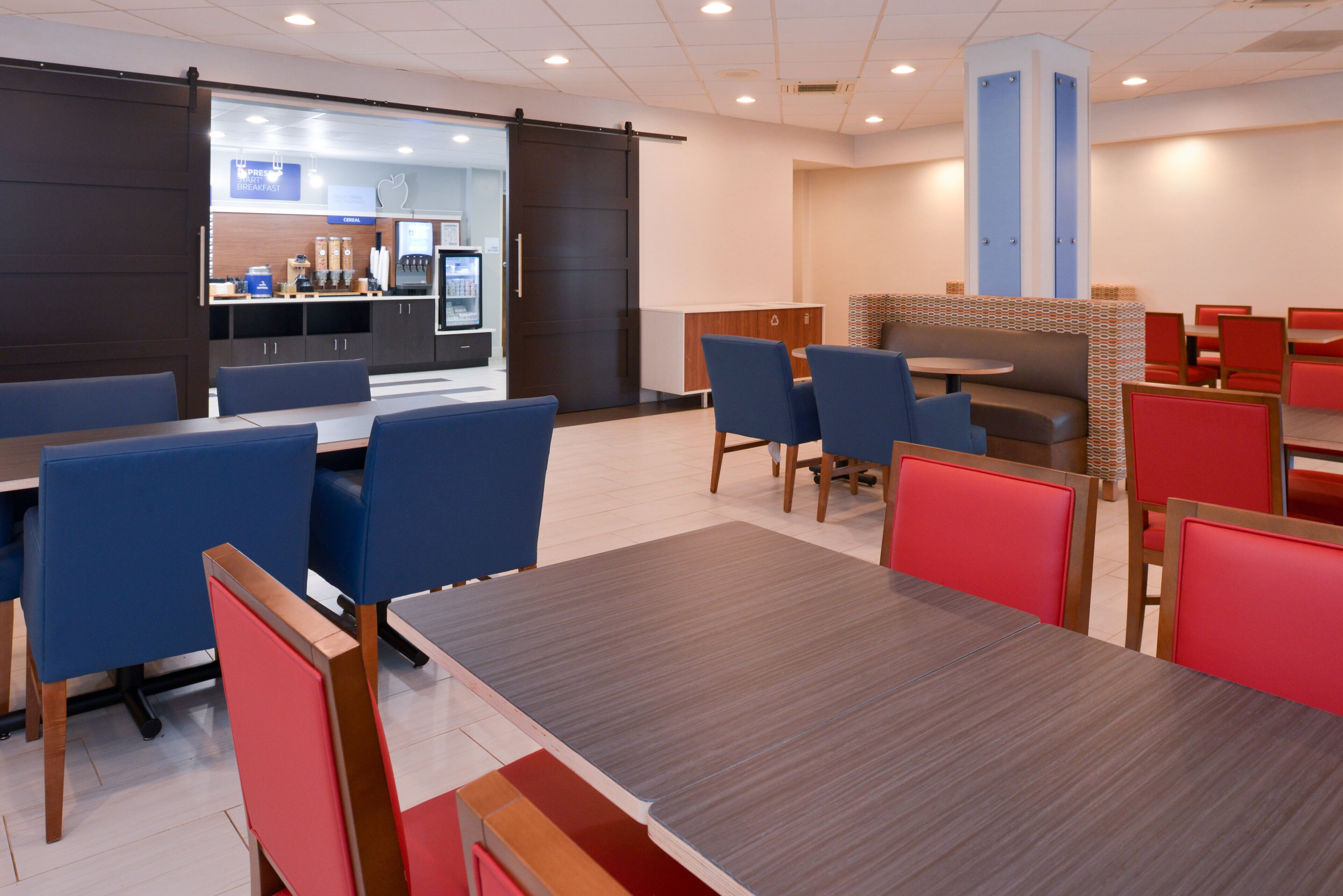 Uma área de refeições moderna, com assentos em cabines azuis e vermelhas, uma mesa central e uma cozinha ao fundo.