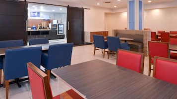 Uma área de refeições moderna, com assentos em cabines azuis e vermelhas, uma mesa central e uma cozinha ao fundo.